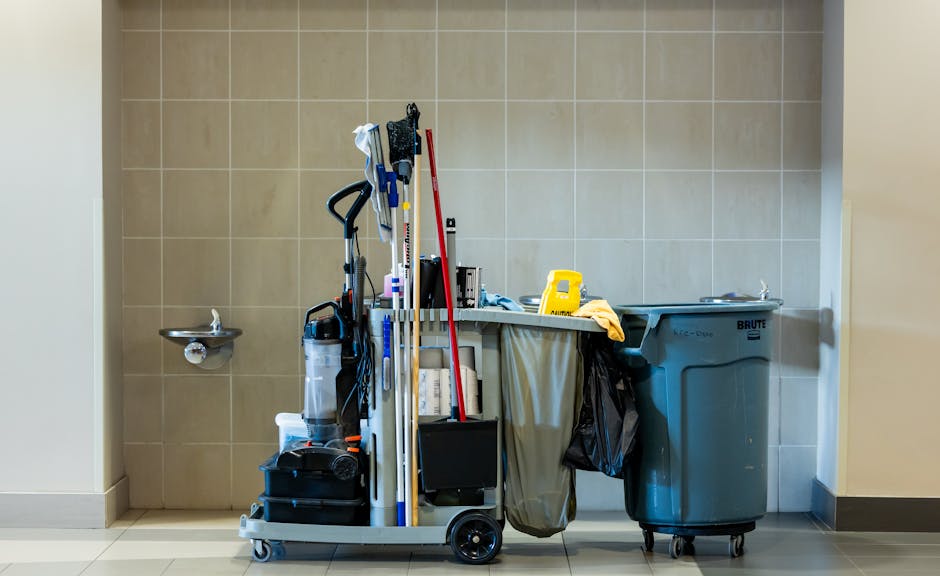 A cleaning trolley filled with various tools and supplies, including mop handles, brushes, and cleaning sprays, positioned next to a large blue waste bin in a tiled room with beige walls. The trolley